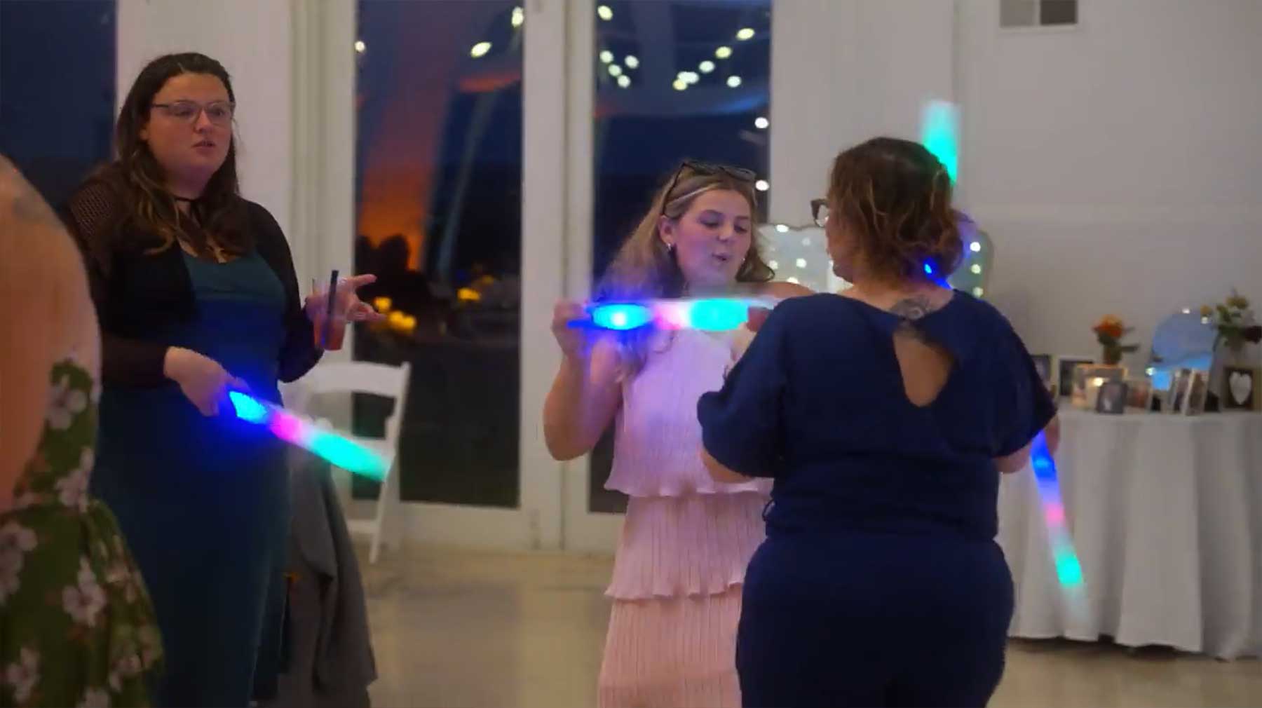 Rotunda-at-Lauxmont-Farms-wedding-guests-dancing Three women dance indoors, holding and waving colorful glowing sticks, with a decorated table and white chairs in the background.