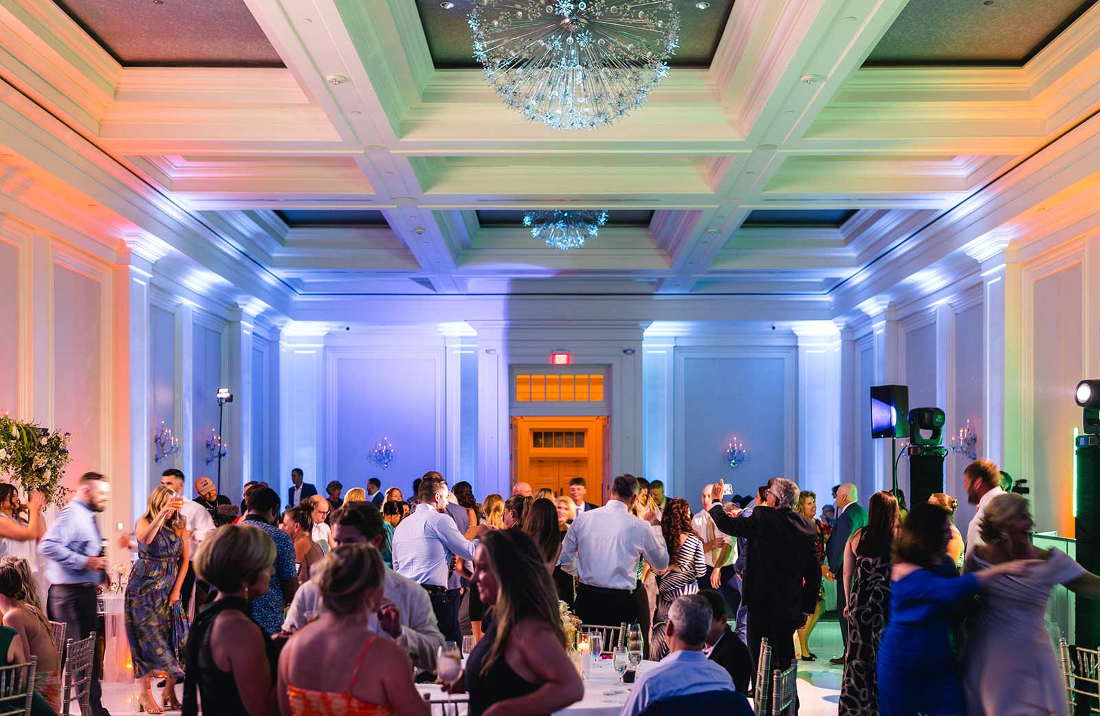 A large group of people attend an indoor event in a brightly lit banquet hall with high ceilings and a chandelier, with some guests standing and others dancing.