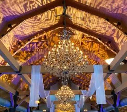 Crystal chandeliers hang from a high wooden ceiling, casting intricate light patterns, with white fabric draped across beams in an event space.