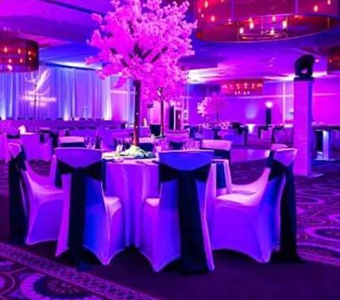 A banquet hall set for an event with round tables, white chairs with dark sashes, and pink-lit artificial trees as centerpieces under purple lighting.