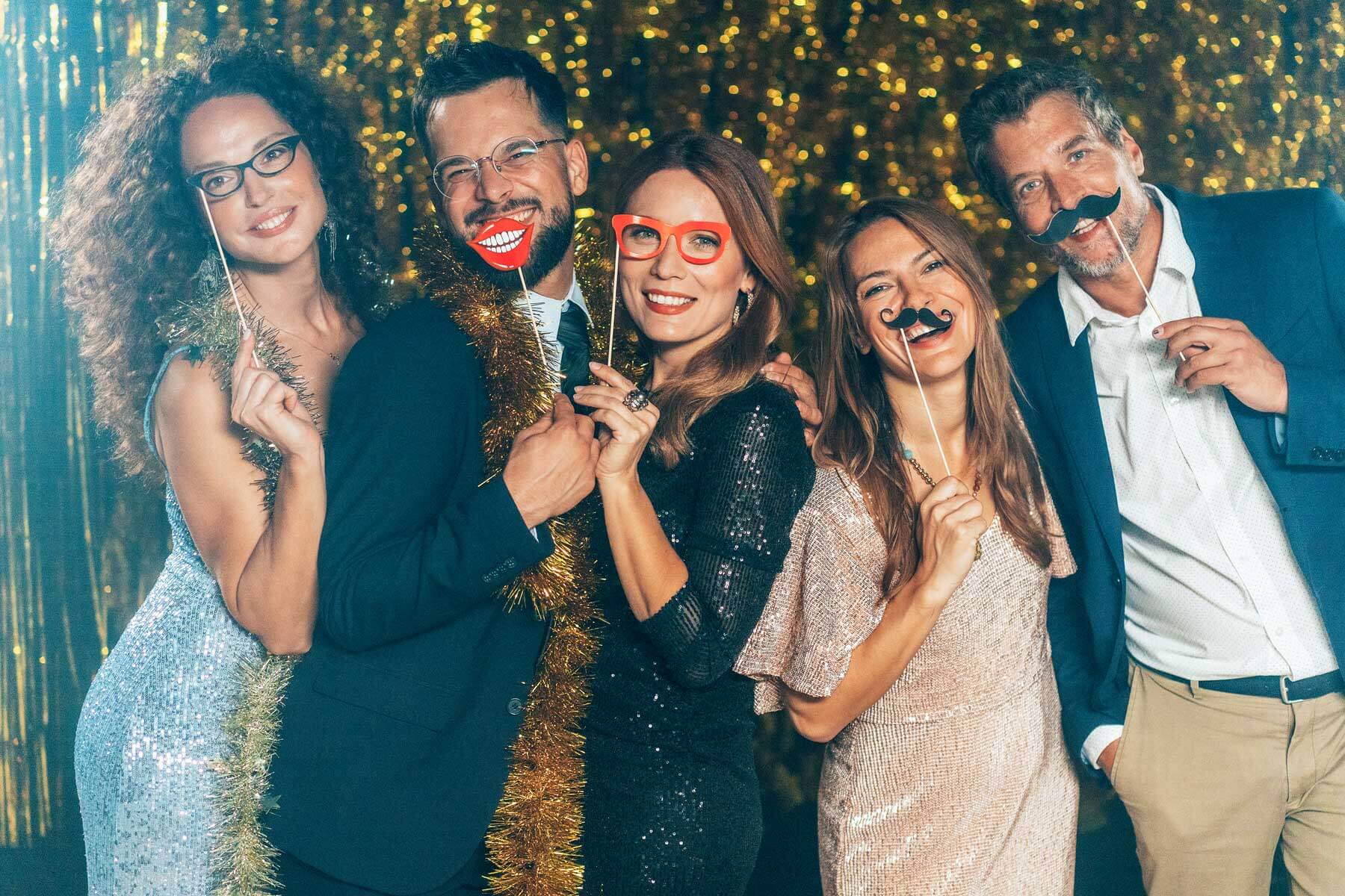 corporate-photo-booth Five people dressed in party attire pose together, holding playful props like fake glasses, lips, and mustaches against a gold shimmering backdrop.
