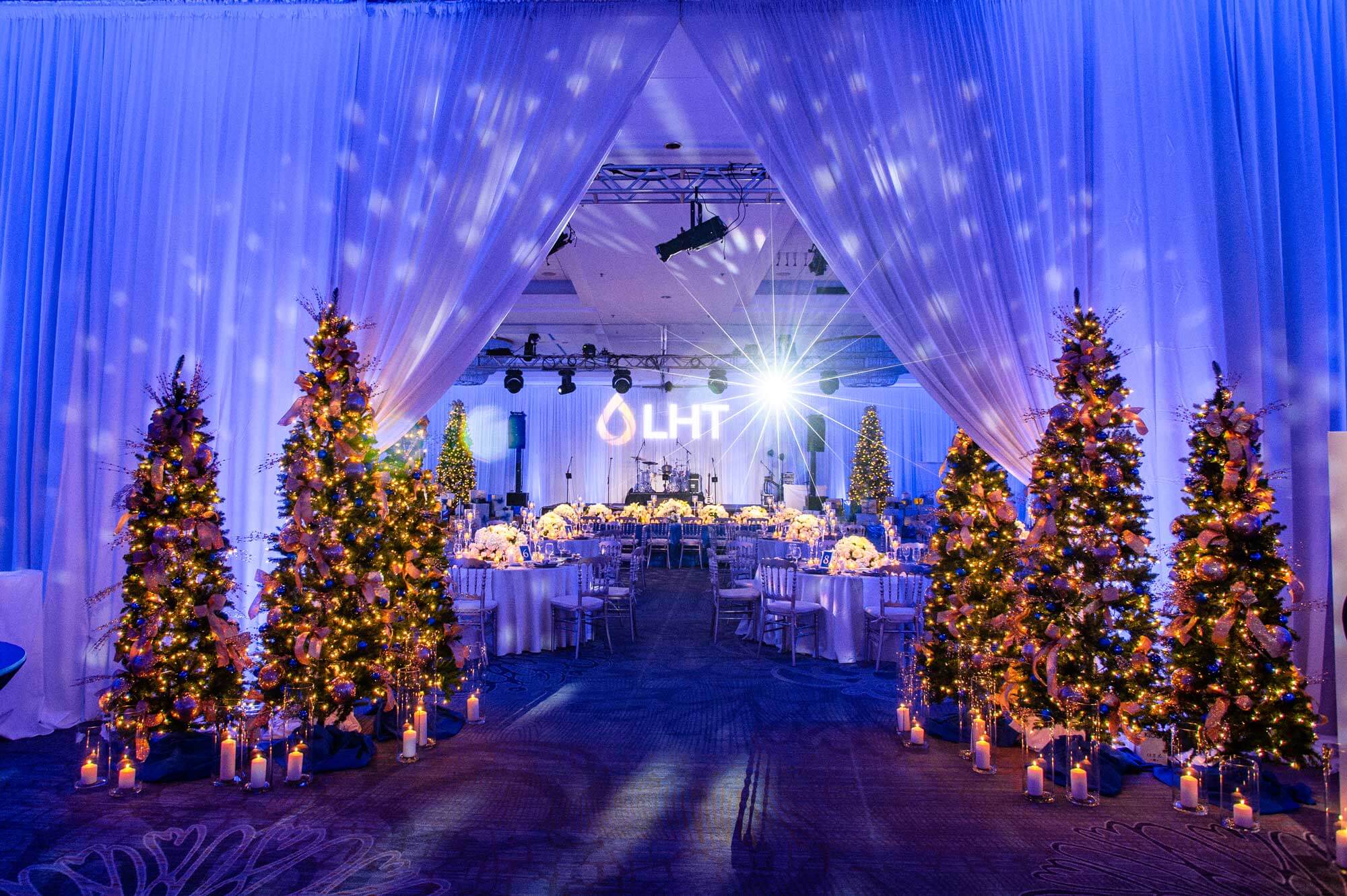 dj-drapery-lighting-Hilton-Harrisburg Ballroom decorated for an event with lit Christmas trees, draped white curtains, and round tables set for dining, illuminated by blue lighting and spotlights.