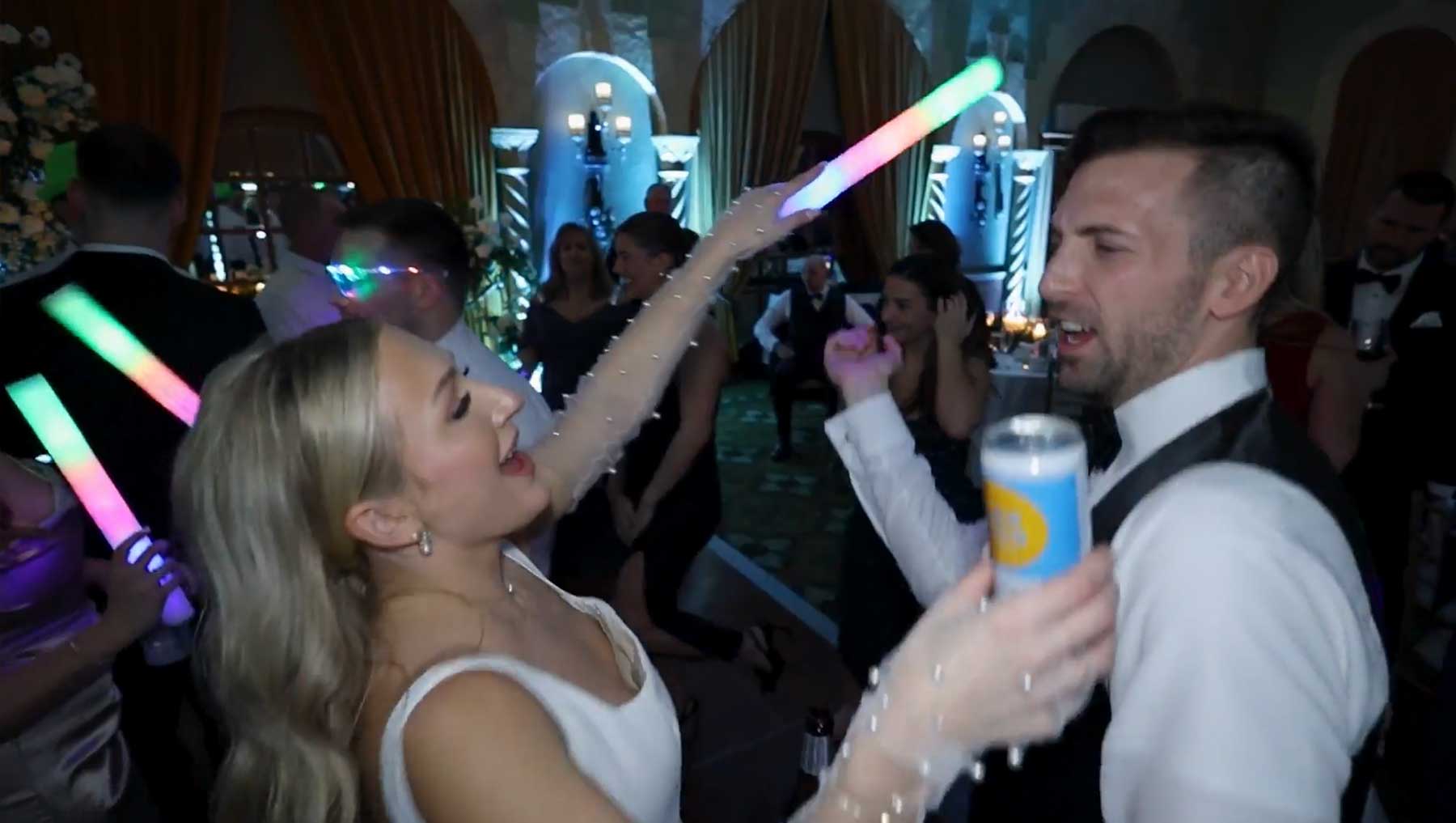 hotel-hershey-wedding-video-bride-and-groom A woman in a white dress and a man in a suit dance at a party, holding colorful light sticks. The man holds a can and other people are dancing in the background.