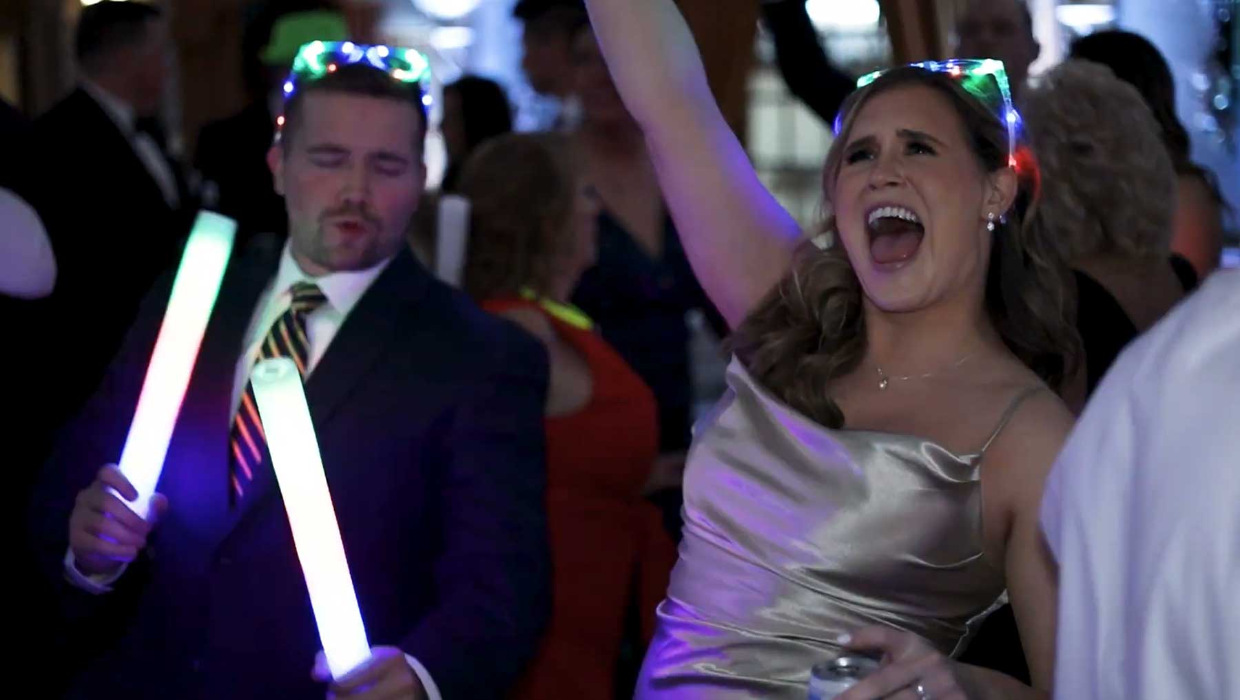 hotel-hershey-wedding-video-dancing-guests Two people in formal wear dance and hold glowing sticks at a lively indoor event, surrounded by other guests.
