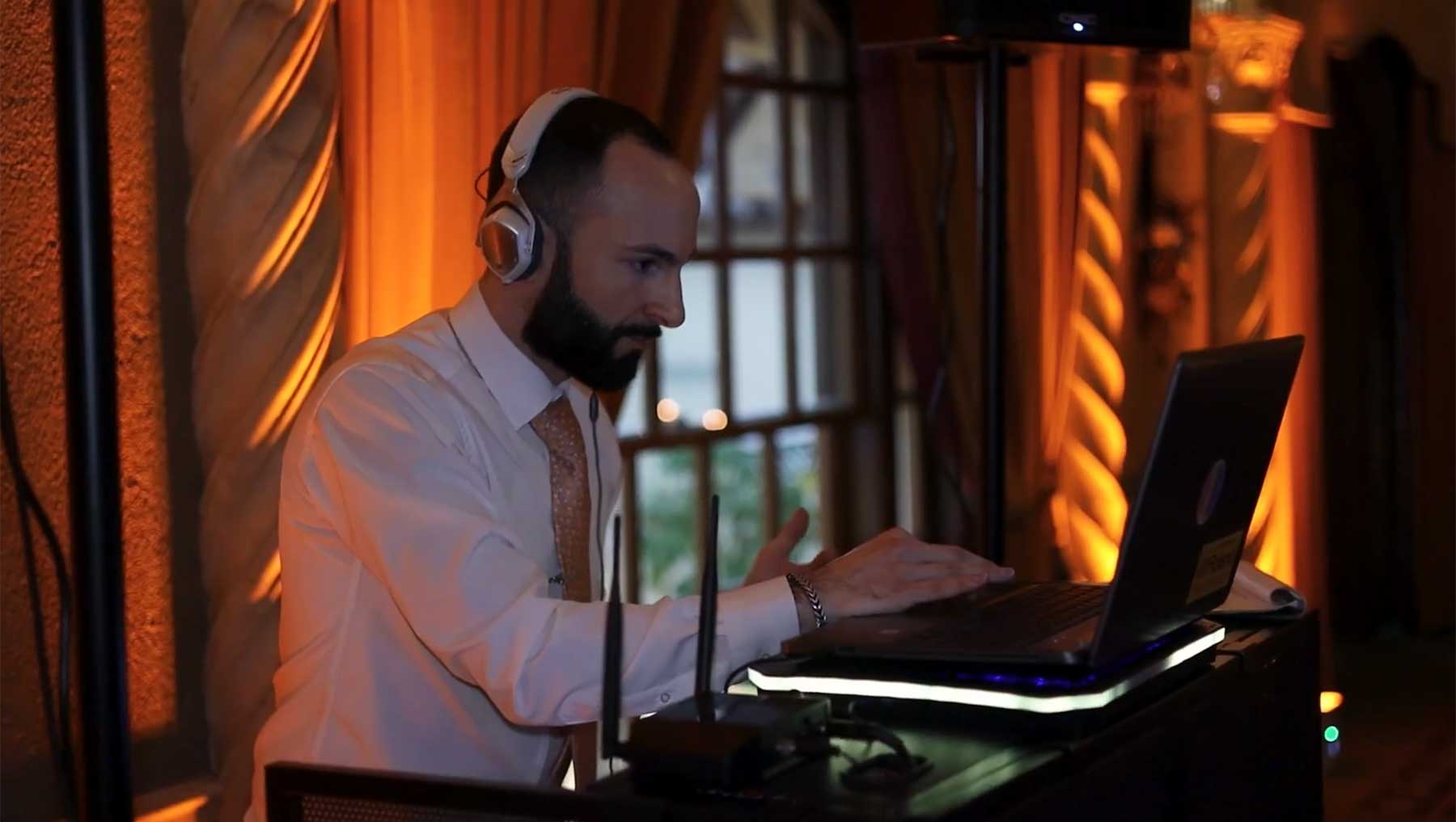 hotel-hershey-wedding-video-dj-josh A man wearing headphones operates a laptop at a DJ booth in a dimly-lit room with warm lighting and decorative columns.