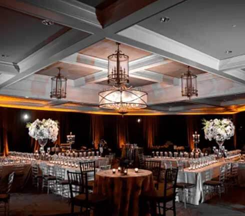 Elegant banquet hall with long dining tables, white floral centerpieces in tall vases, and dim warm lighting under chandelier fixtures.