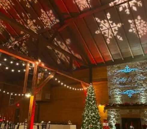 A decorated indoor event space with string lights, a lit Christmas tree, and snowflake patterns projected on the wooden ceiling.