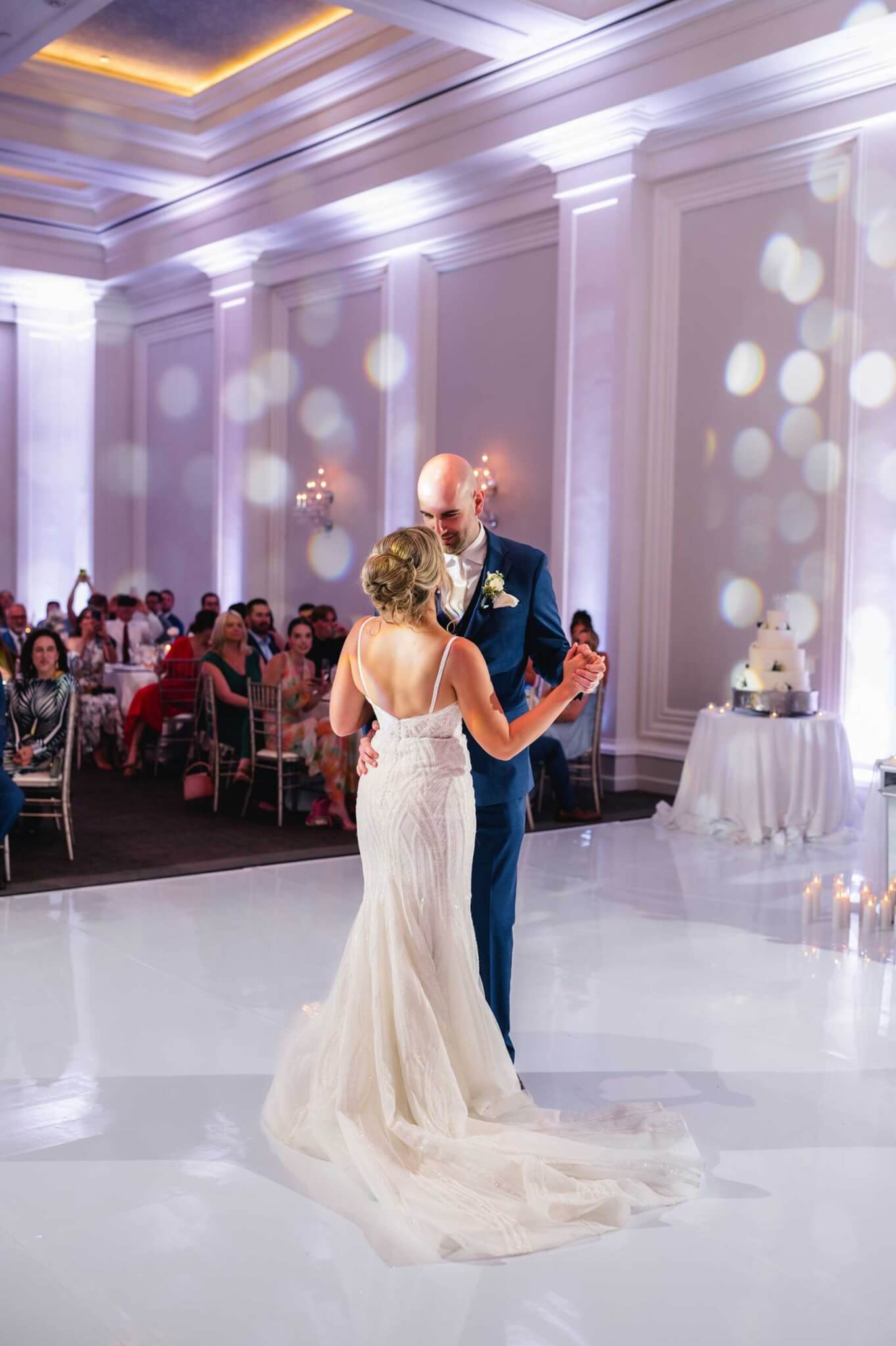 A bride and groom share their first dance on a white floor in an elegant, softly lit ballroom with seated guests watching.