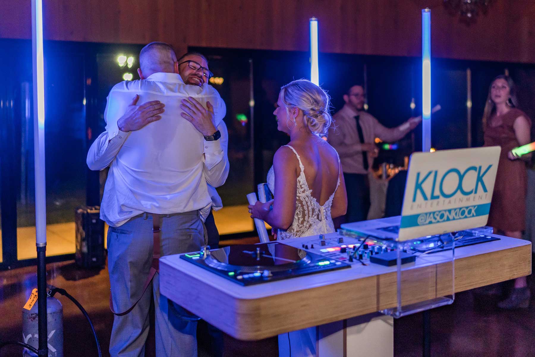 wedding-Historic-Acres-of-Hershey Two men hug near a DJ booth at an indoor event while a woman in a white dress stands nearby. Neon lights and DJ equipment are visible in the foreground.