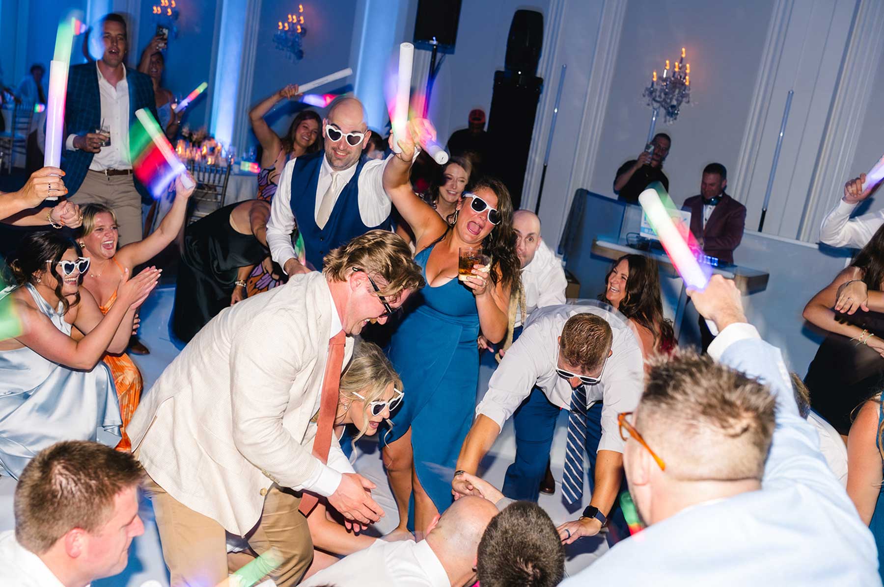 wedding-LF-Ballroom-at-Ellis-Preserve A group of people wearing sunglasses and holding glow sticks dance energetically at a party in a brightly lit indoor venue.