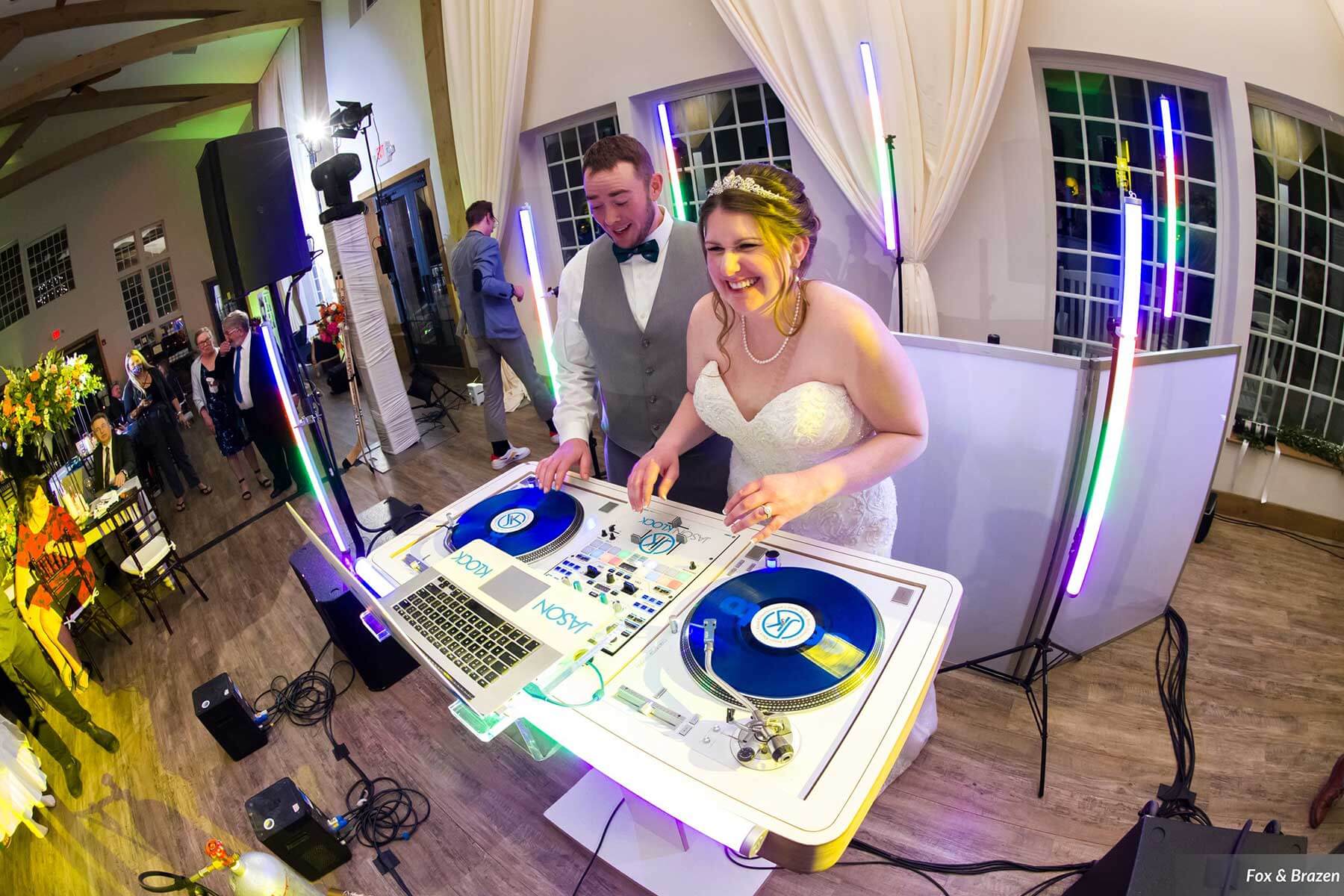 wedding-LF-Stephano-Hopkins-Fox-&-Brazen-FoxBrazen-HopkinsWedding-4785a A bride and groom stand behind DJ turntables at their wedding reception, smiling as they play music together. Neon lights and guests are visible in the background.