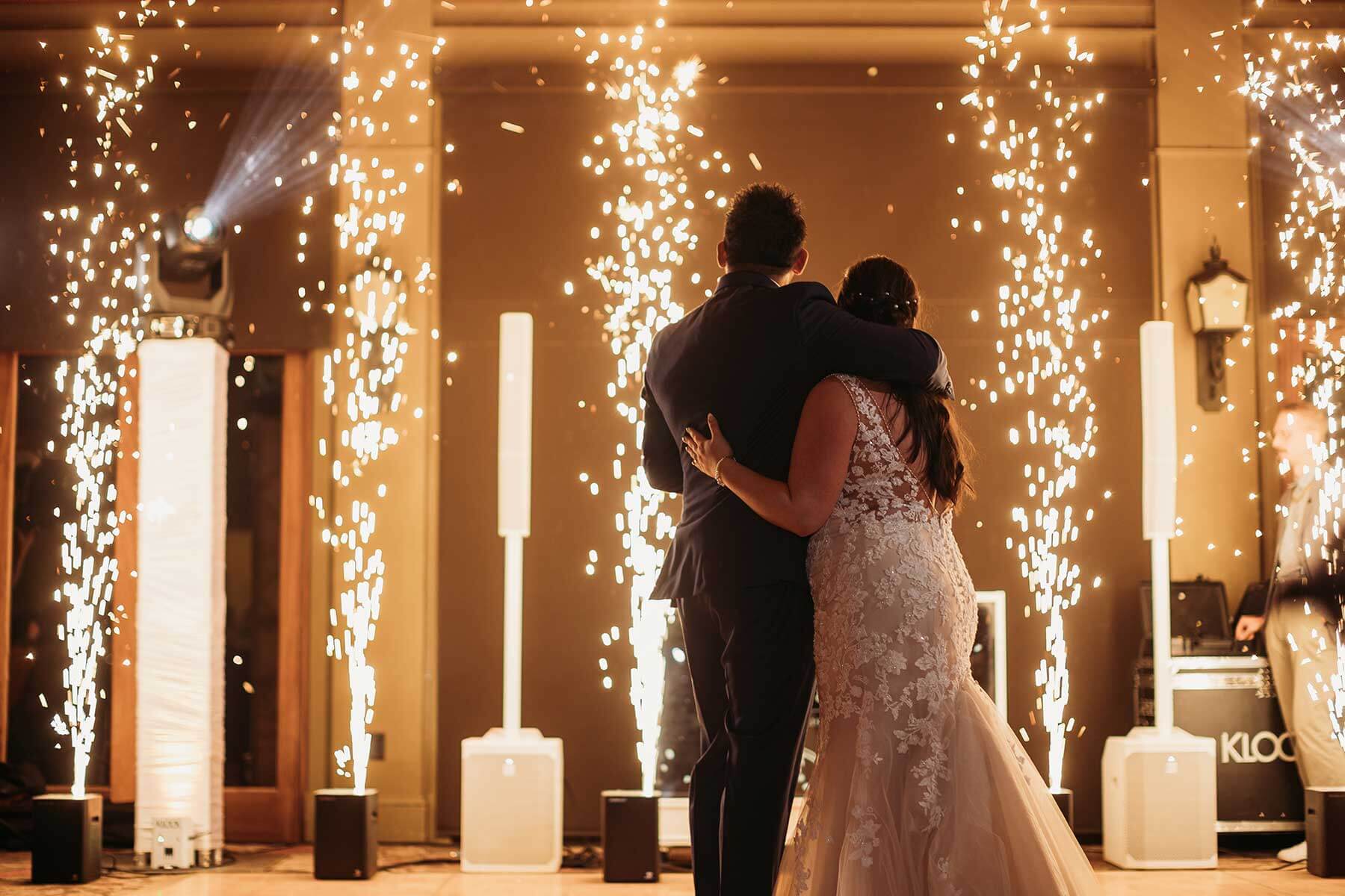 A couple in formal attire stands arm in arm, facing away, as indoor fireworks and sparklers light up the background at an event.