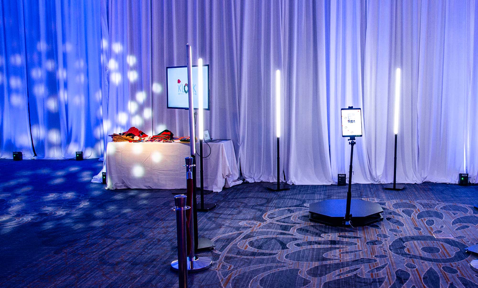 360-photo-booth-setup A photo booth setup with vertical light stands and a tablet on a platform in a room with blue lighting and draped white curtains; a table with props is in the background.