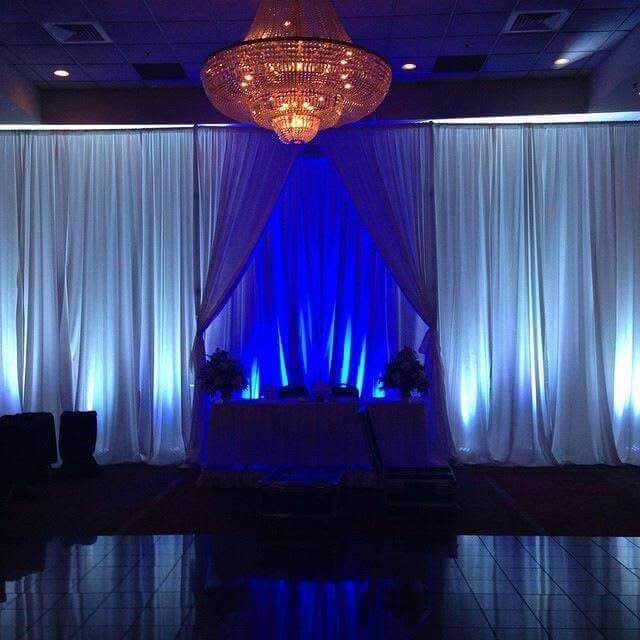 A chandelier hangs above a table set against white drapes illuminated with blue lighting in an event space.