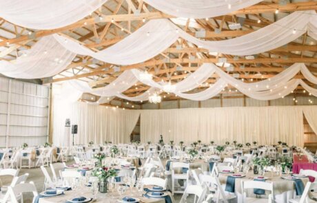 Large event space with white draped fabric on the ceiling, round tables with white chairs, and simple floral centerpieces set for a formal gathering.