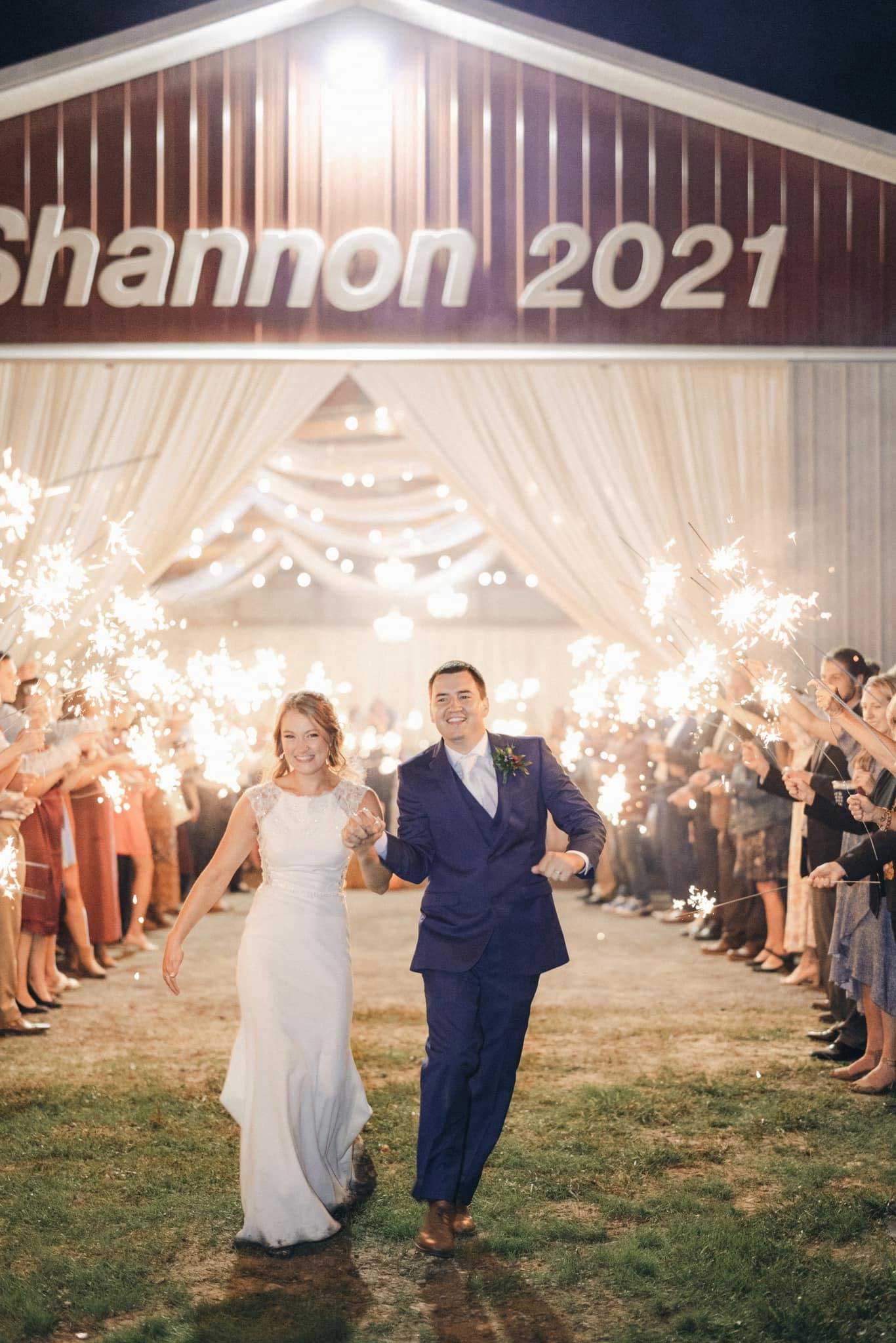 A bride and groom walk hand-in-hand under sparklers, exiting a barn with "Shannon 2021" displayed above the entrance, surrounded by cheering guests at night.