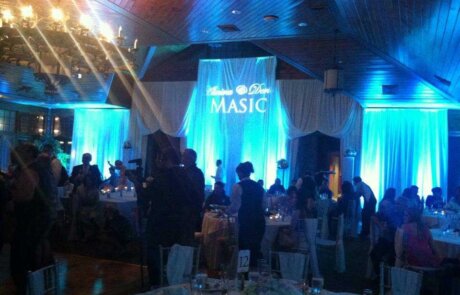 A banquet hall with blue uplighting, round tables set for dinner, and guests mingling; a large sign reads "Masic" above the dance floor.