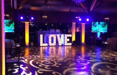 A dimly lit event space with a large illuminated "LOVE" sign, decorative lighting patterns on the floor, and a chandelier overhead.