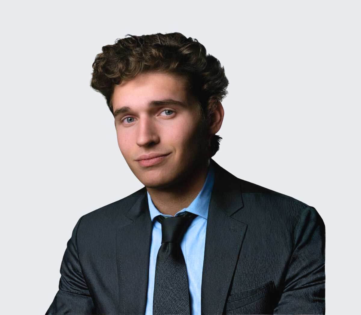 A young man with curly brown hair wearing a dark suit, light blue shirt, and black tie, posed against a plain light background.