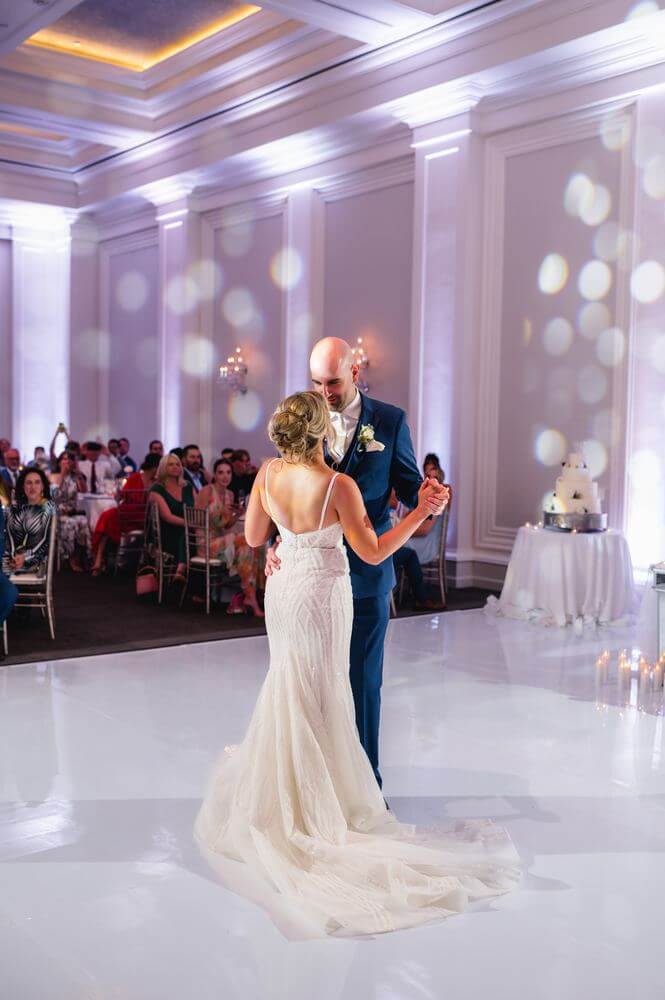 about-room-1 A bride and groom dance together on a glossy white floor in a decorated reception hall, with guests seated and watching in the background.
