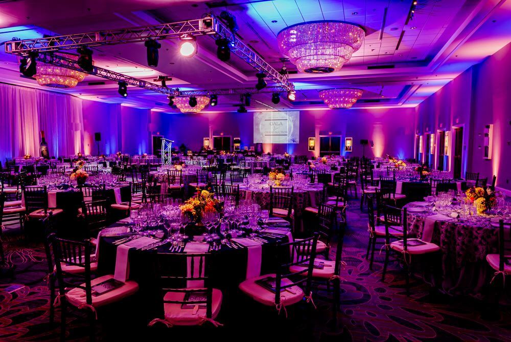 about-room-2 Large banquet hall set up for a formal event with round tables, floral centerpieces, wine glasses, and purple ambient lighting. Chandeliers and a projector screen are visible.