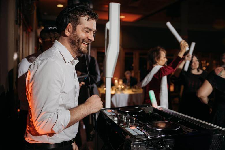 A DJ wearing headphones stands at a mixer, smiling, while people dance and celebrate in a dimly lit event space.