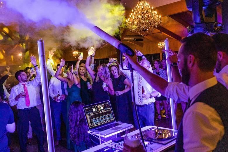 about-slider-2 A DJ performs at a lively indoor party as people dance and raise their hands under colorful lights and fog effects.