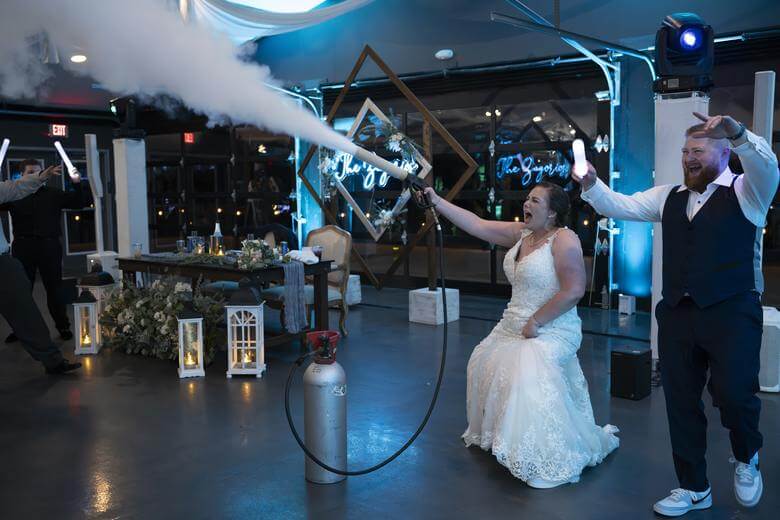 A bride in a wedding dress sprays fog from a canister while guests cheer and hold light sticks at a decorated indoor reception.