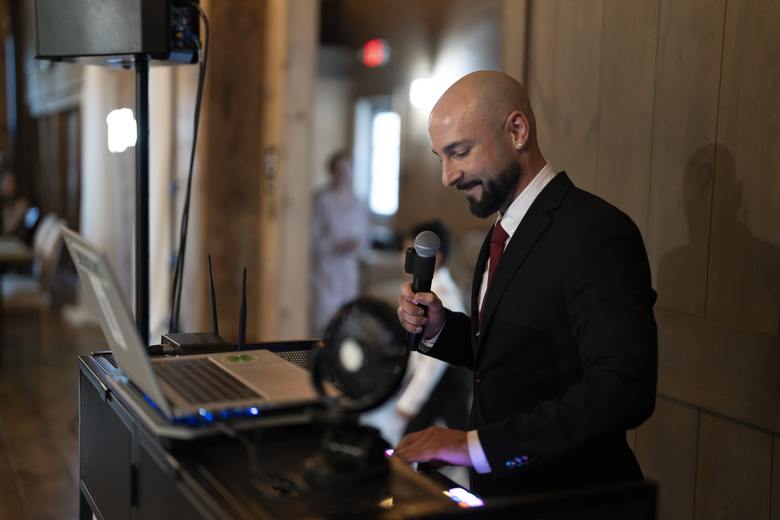 A man in a suit holding a microphone stands at a podium with a laptop and a small fan, preparing to speak in an indoor setting.