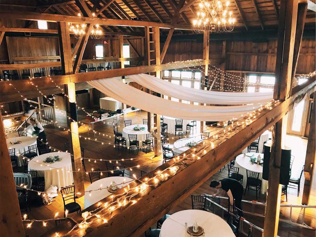 Rustic event venue with round tables, black chairs, white tablecloths, string lights, and draped fabric under wooden beams. A few people are setting up the space.