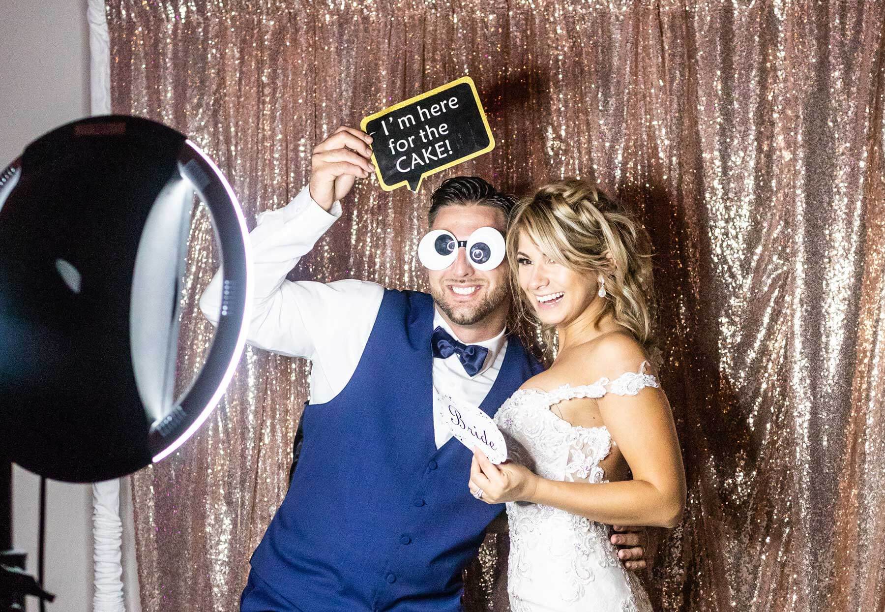 A couple poses in front of a glittery backdrop; the man holds a “I’m here for the cake!” sign and wears oversized googly eye glasses, while the woman wears a white dress and holds a “bride” sign.