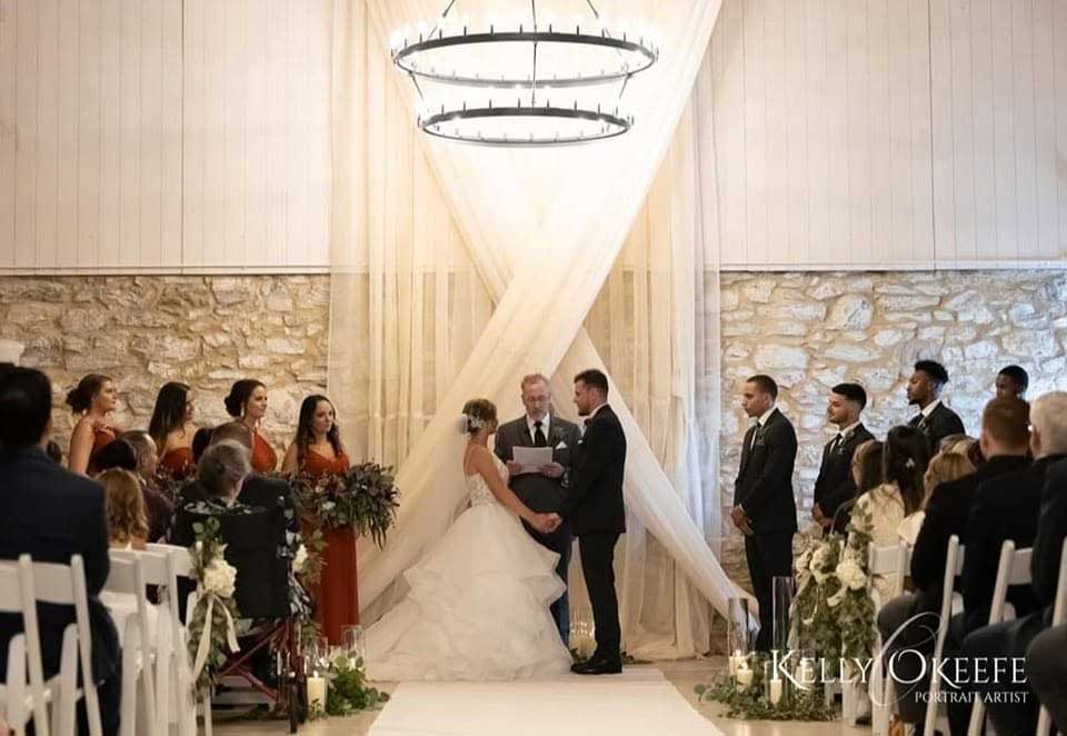 A bride and groom stand at the altar holding hands during their wedding ceremony, surrounded by their wedding party, in a decorated indoor venue with white drapes and stone walls.