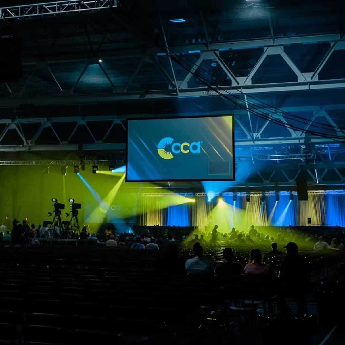 Large indoor event space with a stage, audience seating, blue and yellow lighting, and a screen displaying the CCA logo. Some people are seated, and cameras are set up in the back.