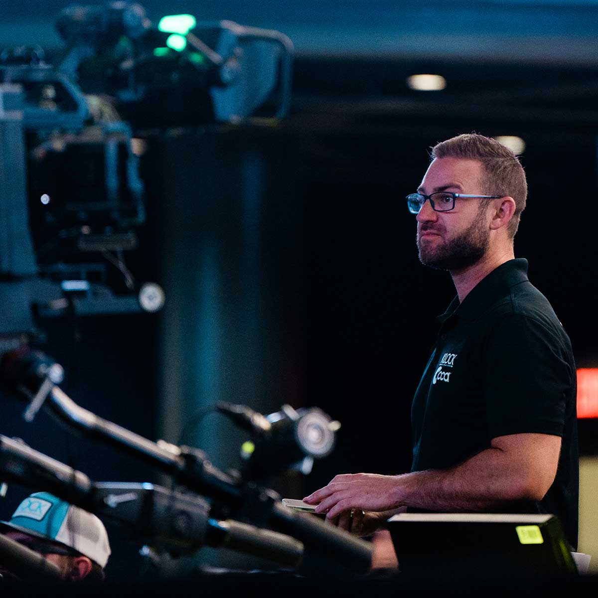 A man with glasses stands at a control panel in a dimly lit room, operating video production equipment.
