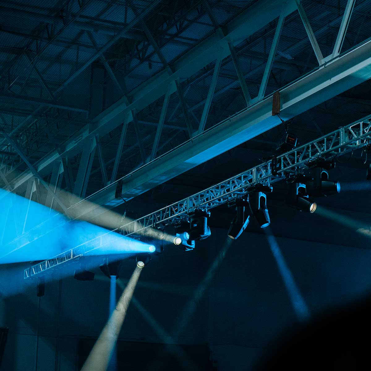 Overhead view of stage lighting equipment and beams of stage lights in a large indoor venue with exposed ceiling beams.