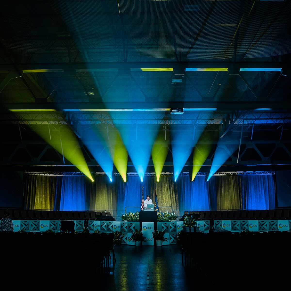 A person stands at a podium on a stage, illuminated by blue and yellow spotlights, in a large indoor venue with rows of empty seats.