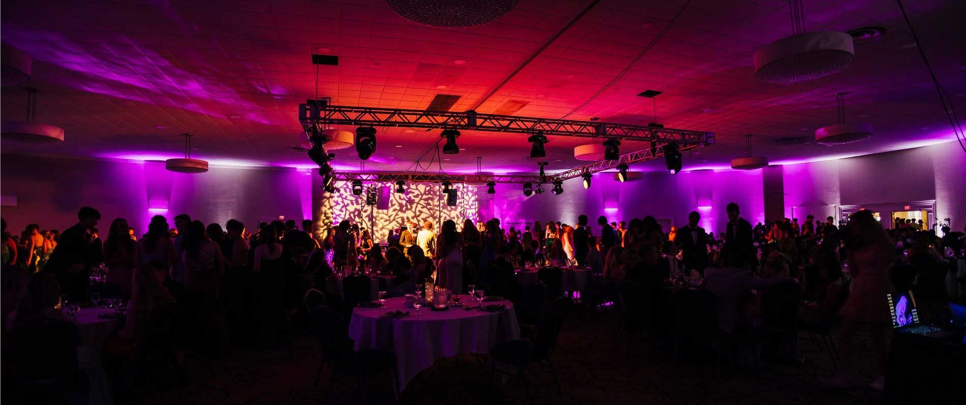 dauphine-prom-party A large indoor event with people mingling and sitting at tables under purple lighting, with a lit stage in the background.