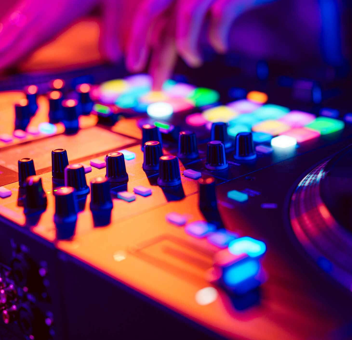 Close-up of a DJ’s hands adjusting controls on a brightly lit mixing console with colorful illuminated buttons and knobs.
