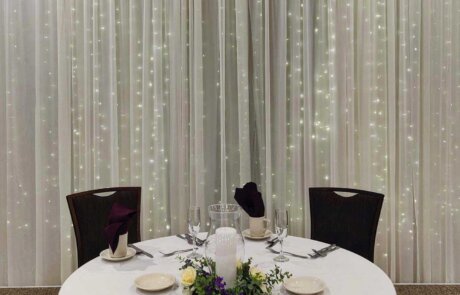 A round table set for two with white linens, glassware, plates, and purple napkins, in front of a backdrop of white curtains with string lights.