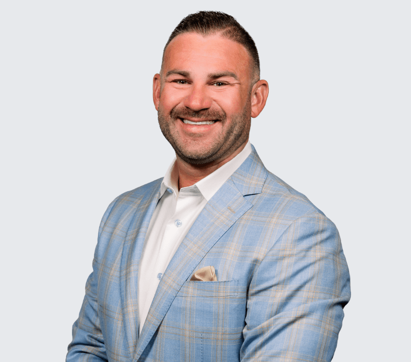 h (1) A man in a light blue plaid suit jacket and white shirt poses smiling with arms folded against a plain, light gray background.