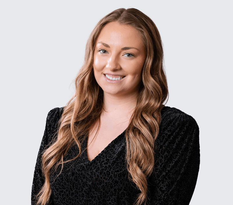 Lexi Sekerak Woman with long wavy light brown hair wearing a black textured top, smiling, posed against a plain light grey background.