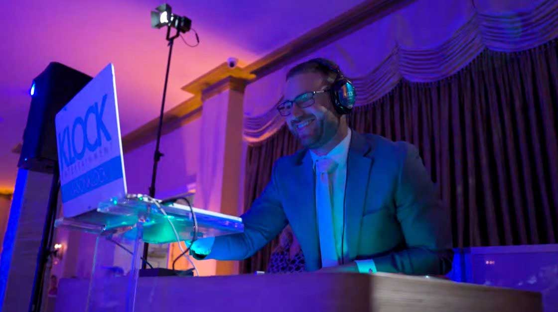 A DJ wearing headphones and a suit is smiling while performing at an event, standing behind a laptop and DJ equipment under purple and blue lighting.