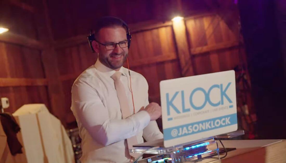 A man wearing headphones and a white dress shirt stands behind a DJ booth labeled "KLOCK" with his hand on the mixer, smiling.
