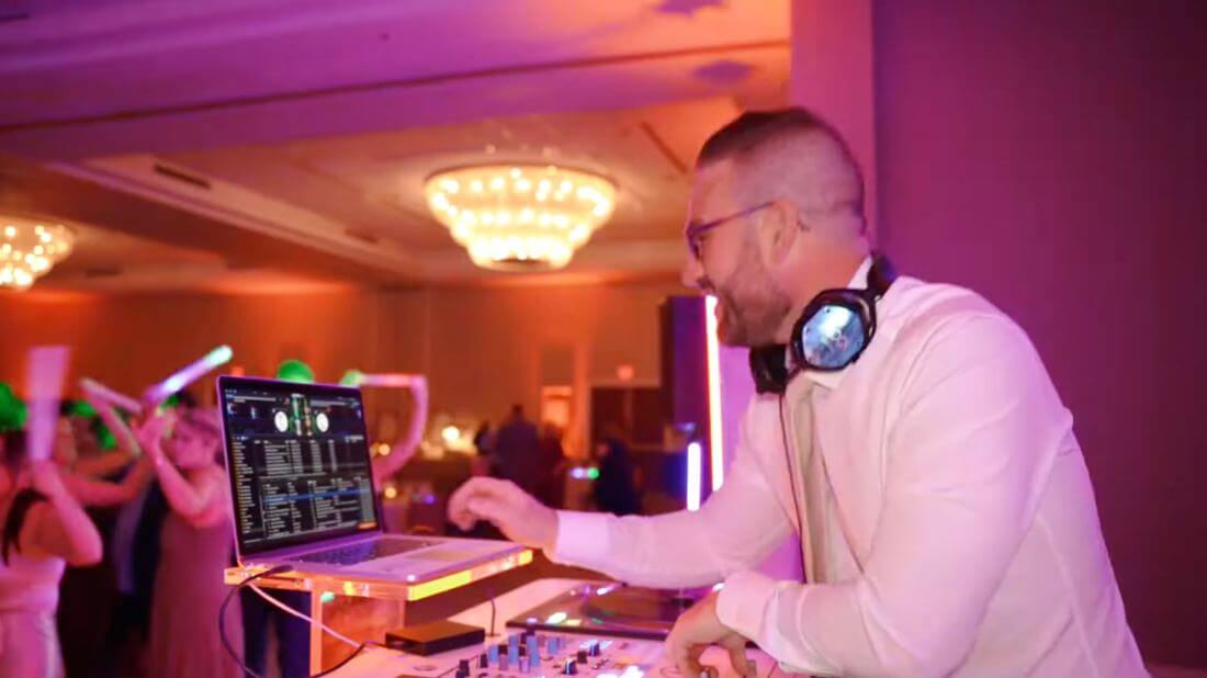 A DJ in a white shirt and headphones stands at a mixing console with a laptop, performing at an indoor event with people dancing in the background.