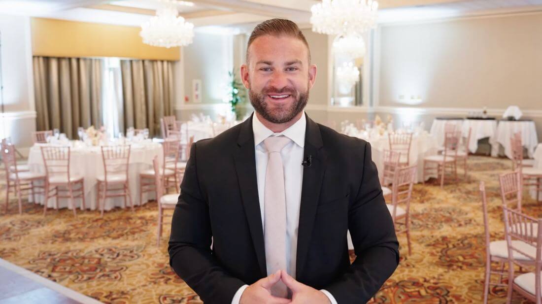 A man in a suit stands smiling in a decorated banquet hall with round tables and chandeliers.