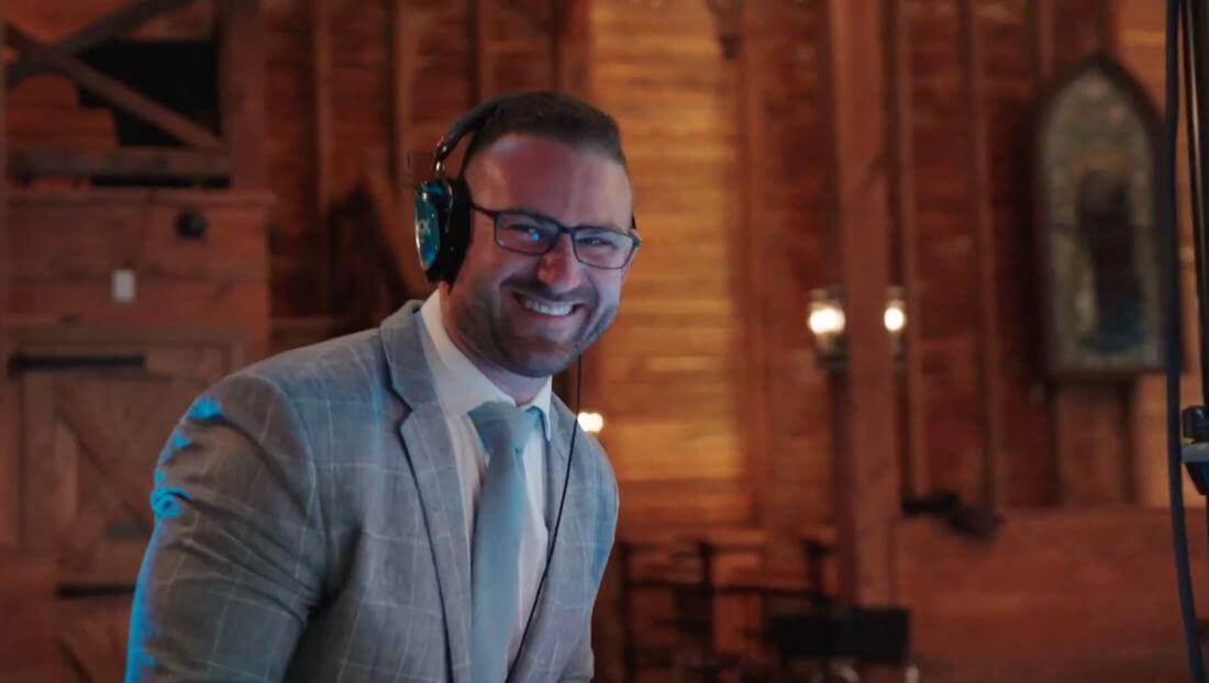 A man in a gray suit and tie, wearing headphones and glasses, smiles while standing indoors in a warmly lit, wooden venue.