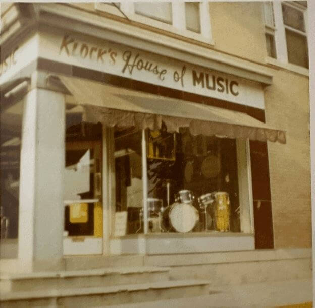 Street-level view of a storefront labeled "Klock's House of Music," displaying a drum set and other musical instruments in the front window.