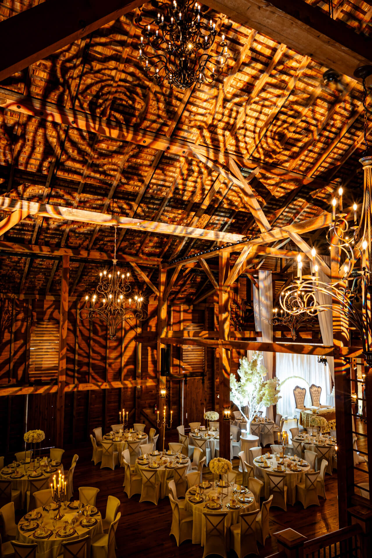 klockstarbarn8.24-3921 Elegant wedding reception setup in a rustic barn with round tables, white floral centerpieces, and dramatic lighting casting patterns on the wooden ceiling and walls.