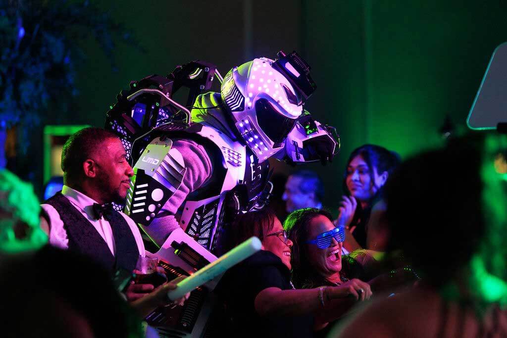 A person in a futuristic LED robot costume stands among a group of people at a lively indoor event, with colorful lights illuminating the scene.