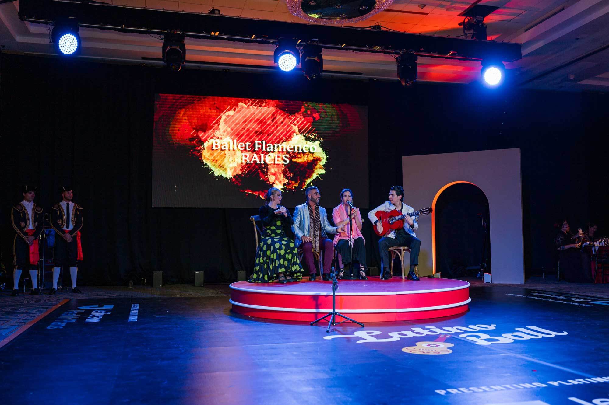Four performers sit on a stage under spotlights, with a guitarist on the right. A screen behind them reads “Ballet Flamenco Raíces.” Two guards stand at the left edge of the stage.